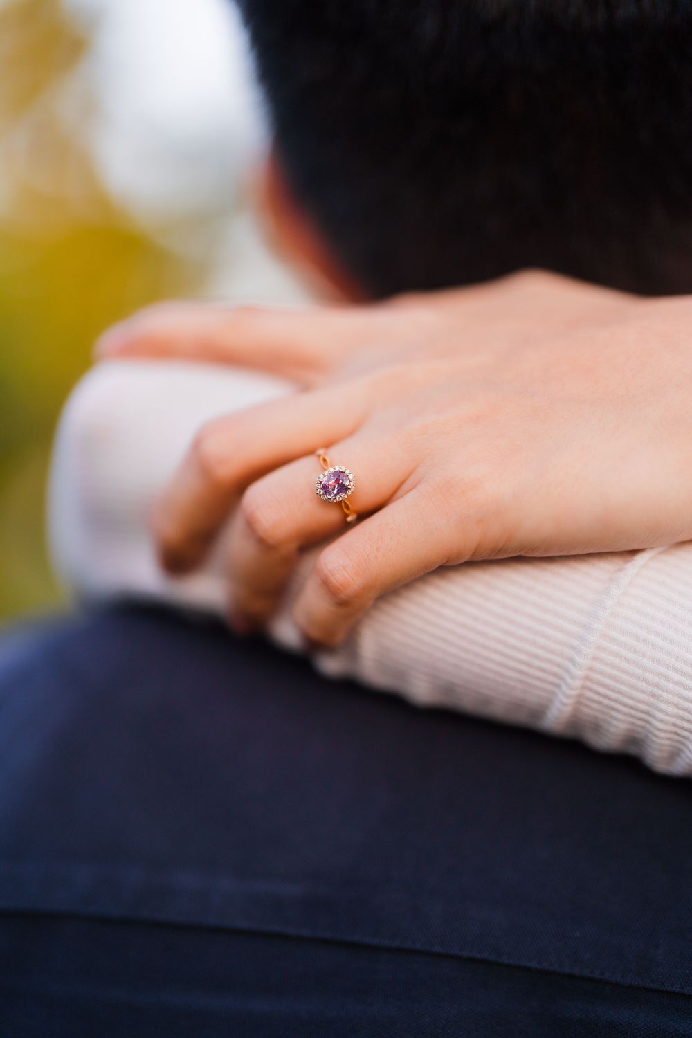 Close-up of a delicate engagement ring with a purple gemstone worn on a hand against a soft background.