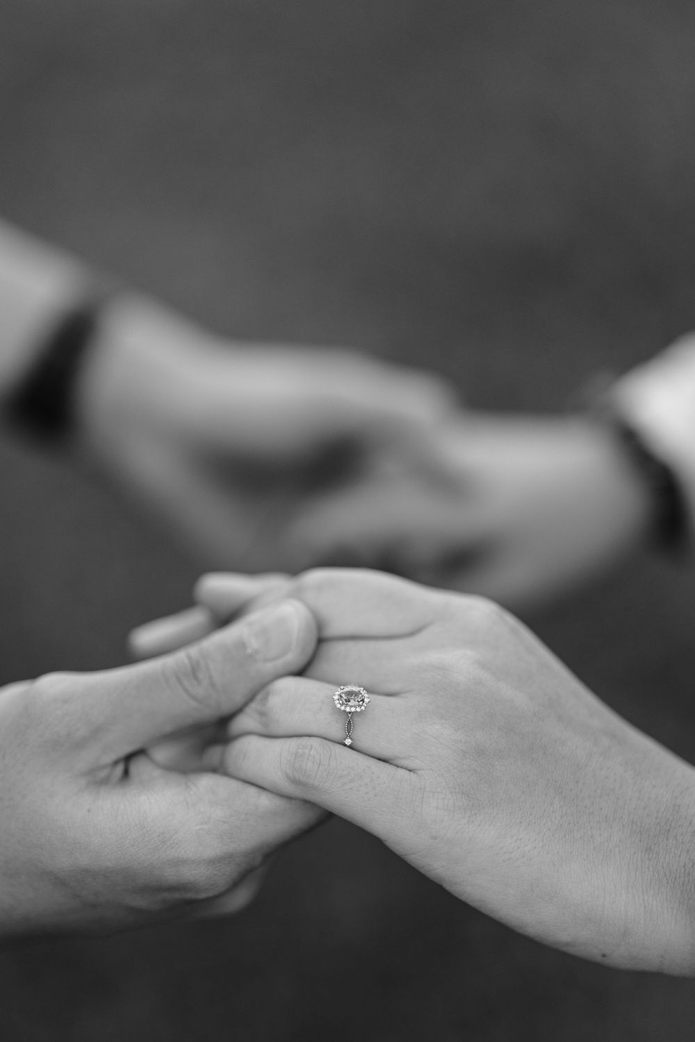 Black and white close-up of hands with engagement ring being placed on finger.