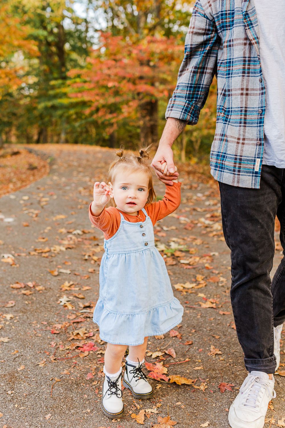 Small child in a denim dress holds hands with adult on fall path covered in leaves.