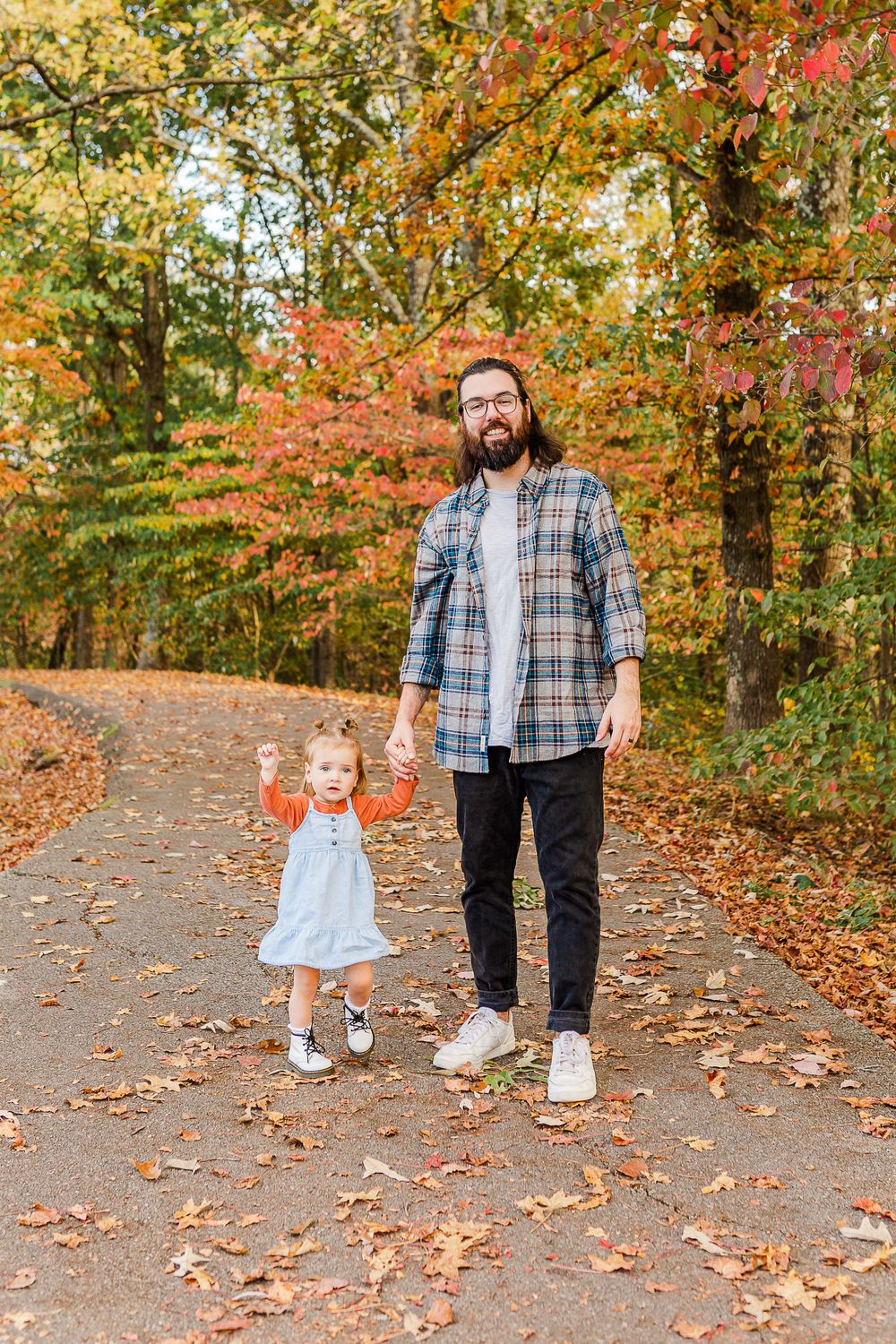 A parent and child walk hand-in-hand down a leaf-covered autumn path.