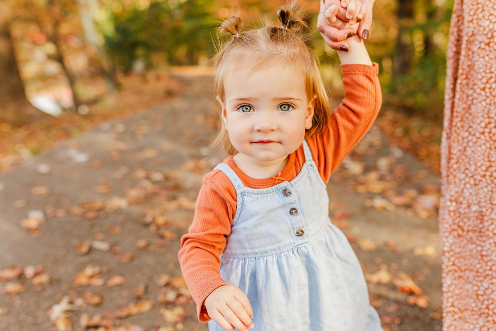 Toddler in orange and blue outfit poses on leaf-covered autumn path.