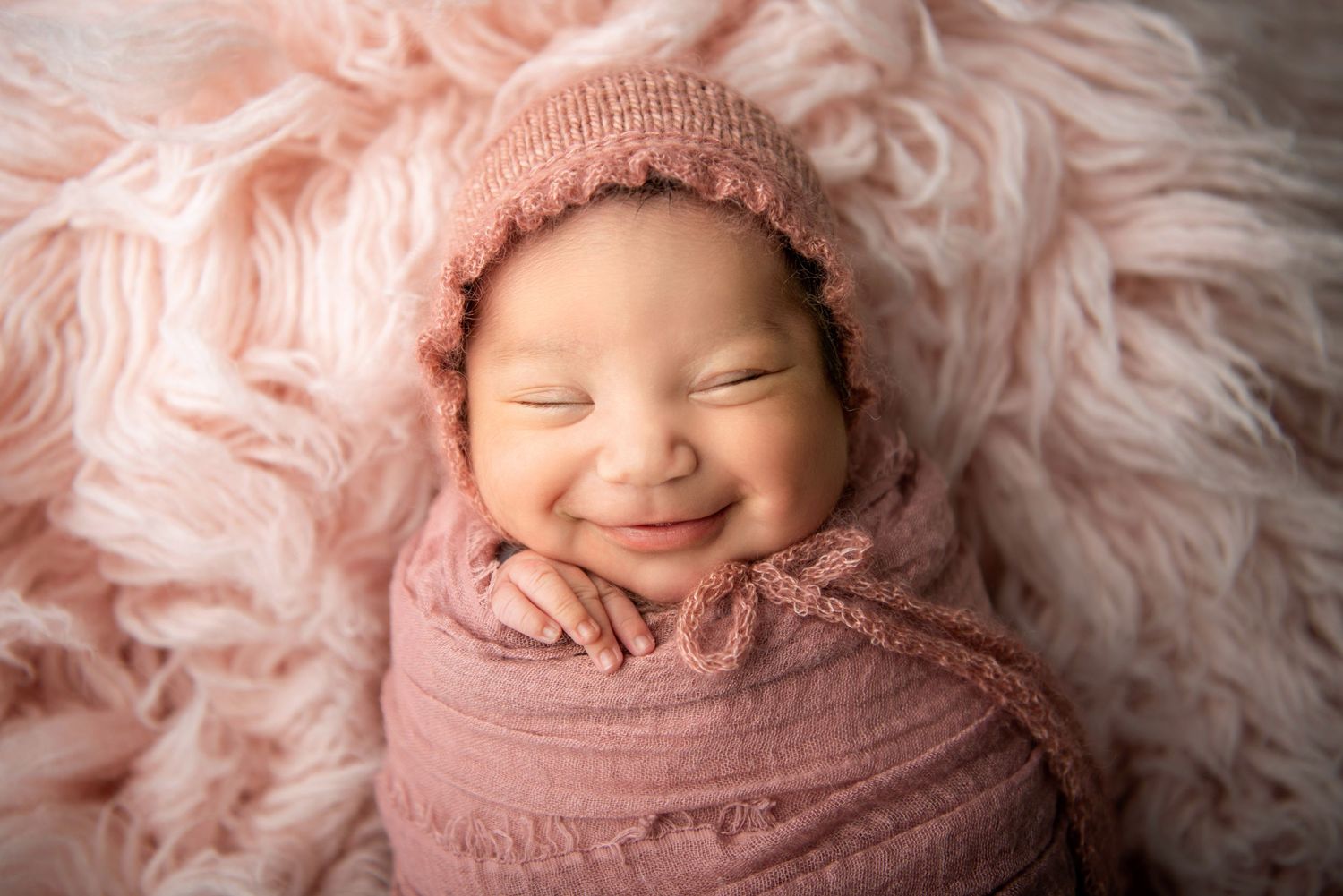 Newborn swaddled in pink wrap with knit bonnet smiling while laying on fluffy pink backdrop.