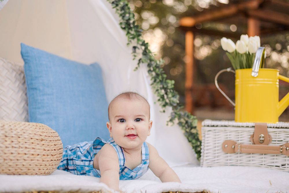 Baby wearing blue romper sits on white blanket with greenery and yellow watering can in background.