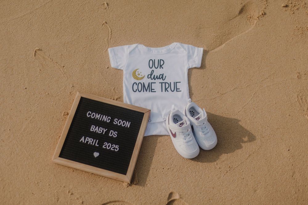 Baby announcement onesie and shoes on sand with chalkboard sign reading Coming Soon Baby 05 April 2025.