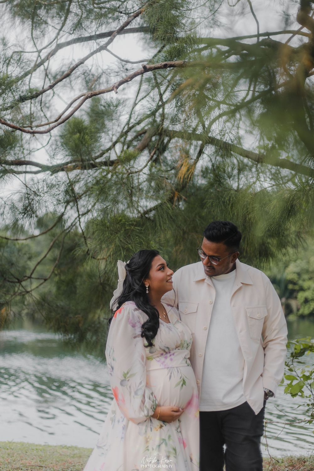 Pregnant couple smiling at each other outdoors near a lake, surrounded by pine trees, woman in floral dress.