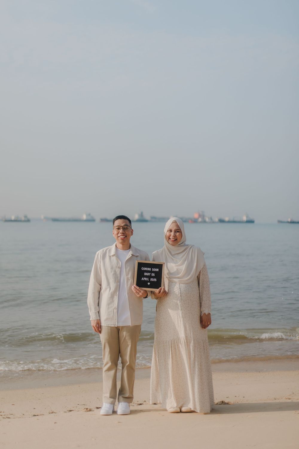 Couple holding a letter board sign on a sandy beach, woman in hijab and white dress, man in beige outfit.