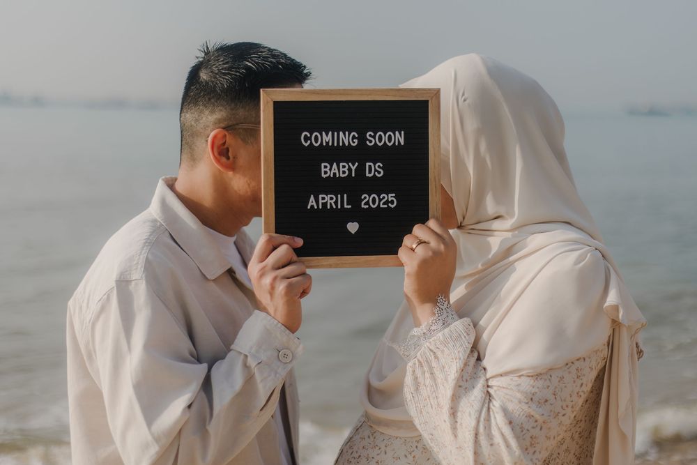 Couple holding a letter board announcing Baby DS coming April 2025, standing on a beach in elegant attire.
