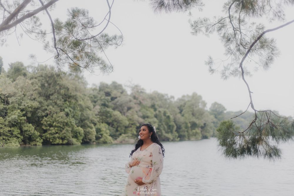 Pregnant woman in lace dress standing by a serene river surrounded by lush green trees on a misty day.