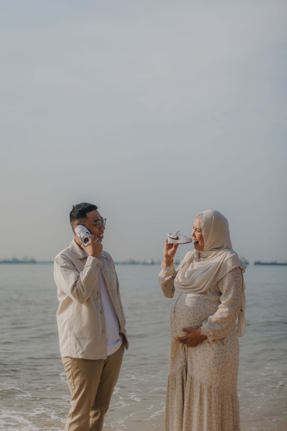 A pregnant Muslim woman in hijab and her husband photograph each other with cameras on a beach.