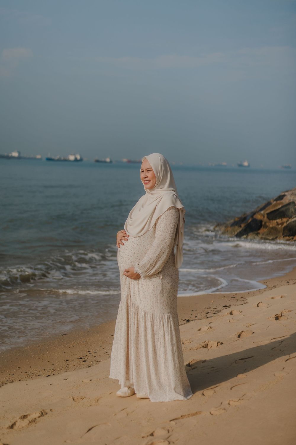 Pregnant woman in white hijab and dress cradling baby bump on sandy beach with ocean waves behind her.