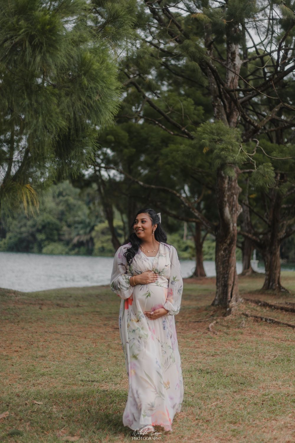 Pregnant woman in floral maxi dress cradling baby bump outdoors near lake surrounded by lush green trees.