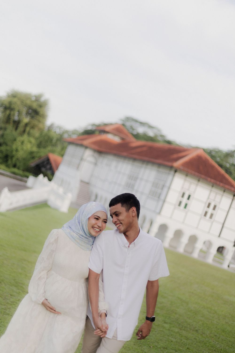 A Muslim couple in white attire poses outdoors on green grass, with a historic white building in the background.