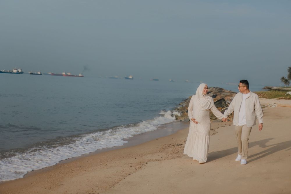 Couple in white outfits walking hand in hand on a sandy beach with calm ocean waves and ships in the background.