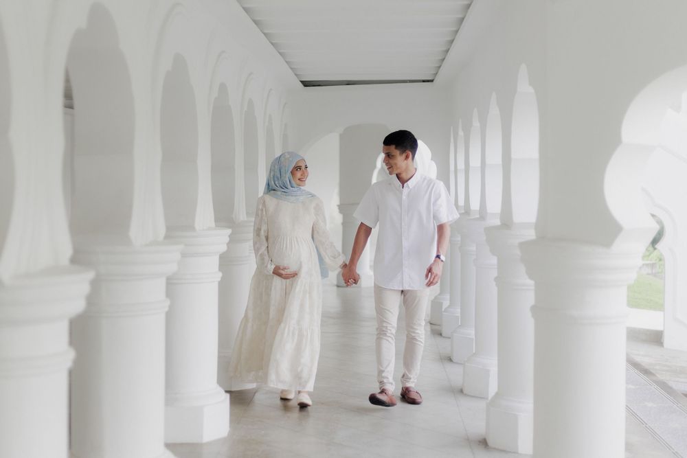Muslim couple in white attire holding hands walking through elegant white arched corridor.