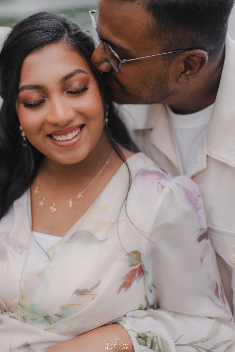 A couple shares a tender moment, man kissing womans cheek as she smiles, both dressed in elegant light outfits.