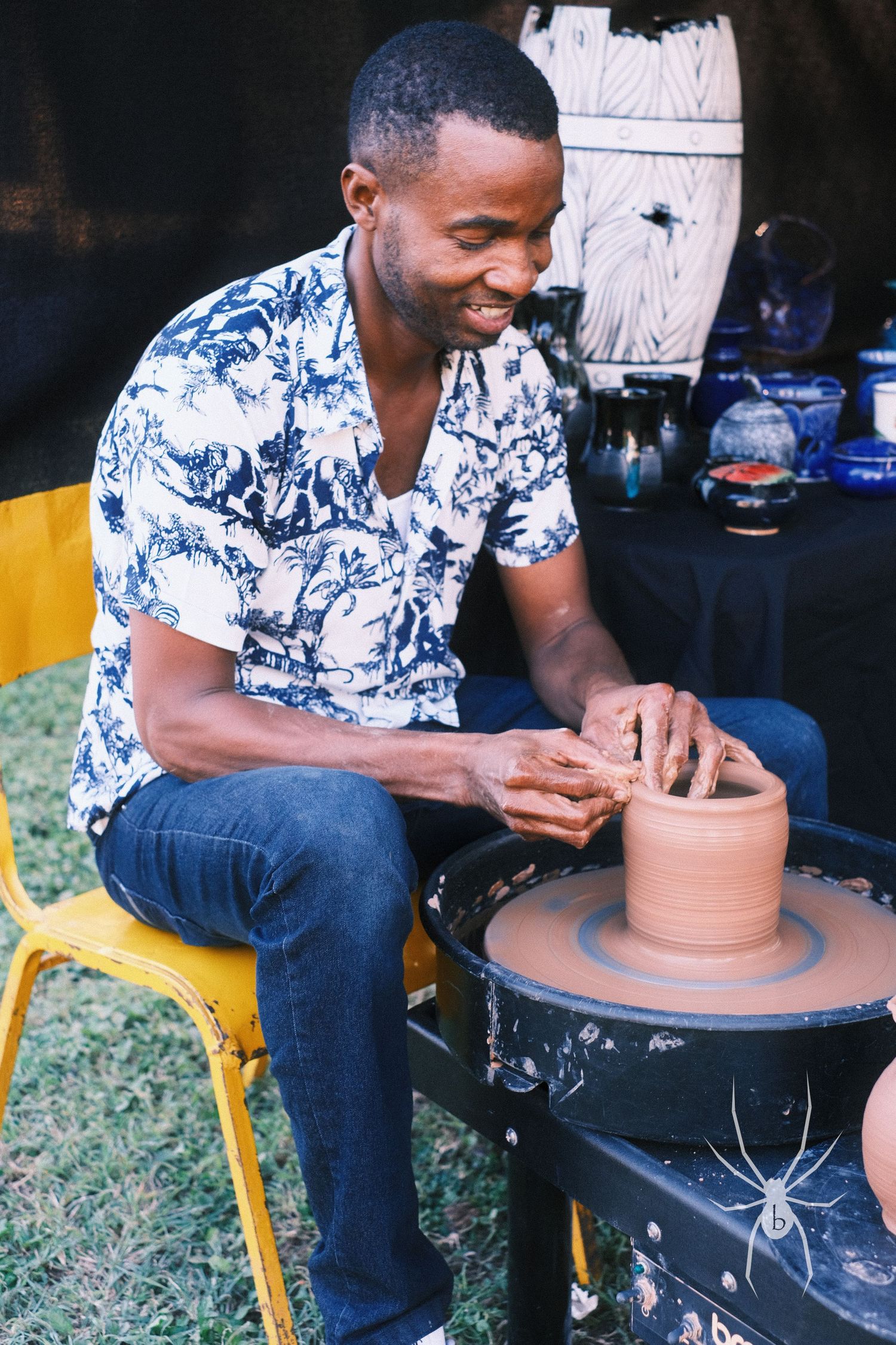 Artist crafting pottery on a wheel while wearing a blue and white floral shirt.