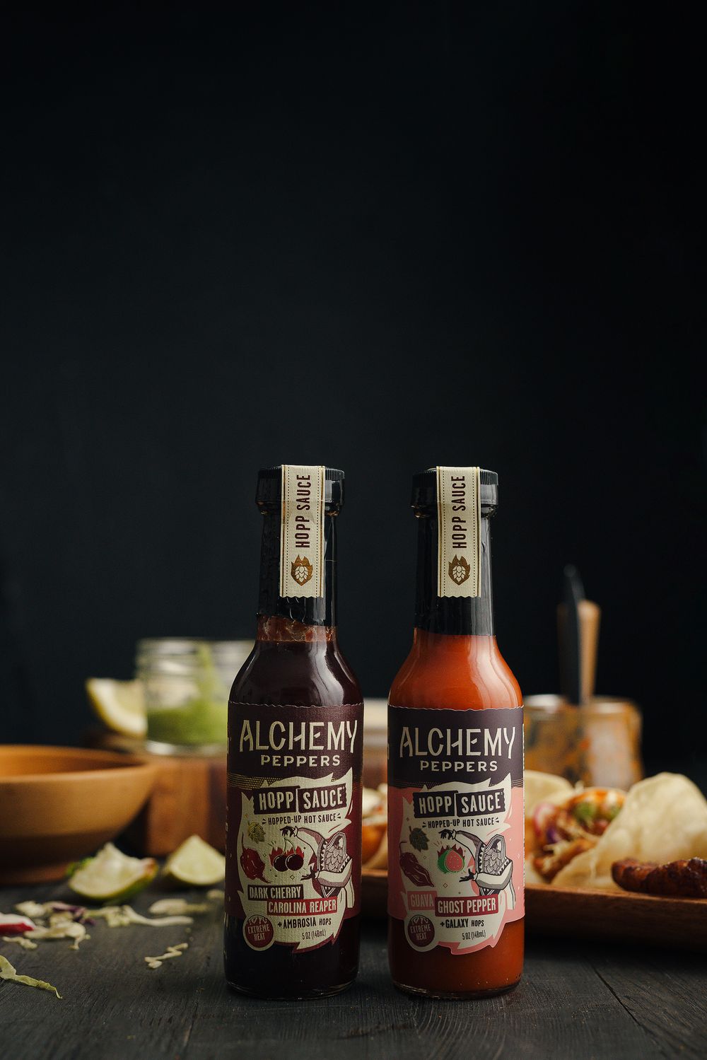 Two bottles of Alchemy brand sauces displayed on a dark wooden surface with lime wedges and food in the background.