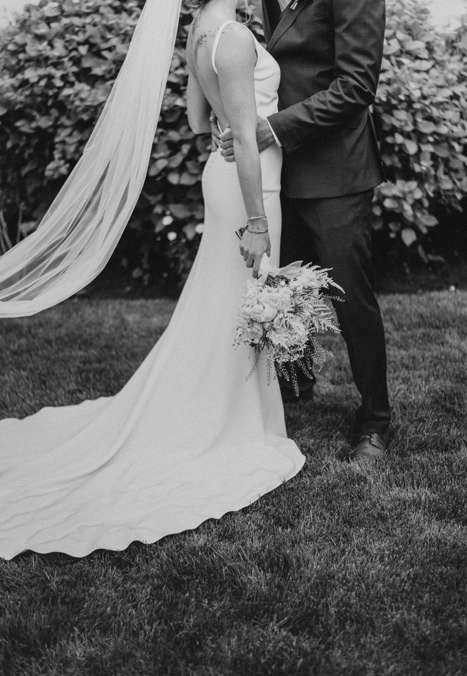 Black and white detail shot of wedding dress train and bouquet against garden backdrop.