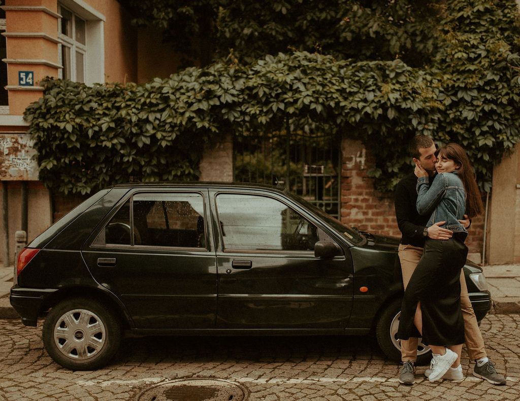 A couple stands close together against a brick wall with trees and parked cars in the background.