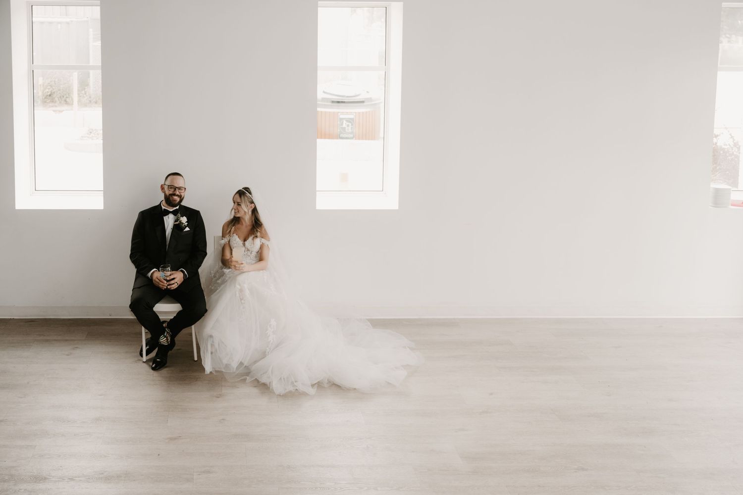 A bride and groom sit together against a white wall in an elegant, minimalist setting.