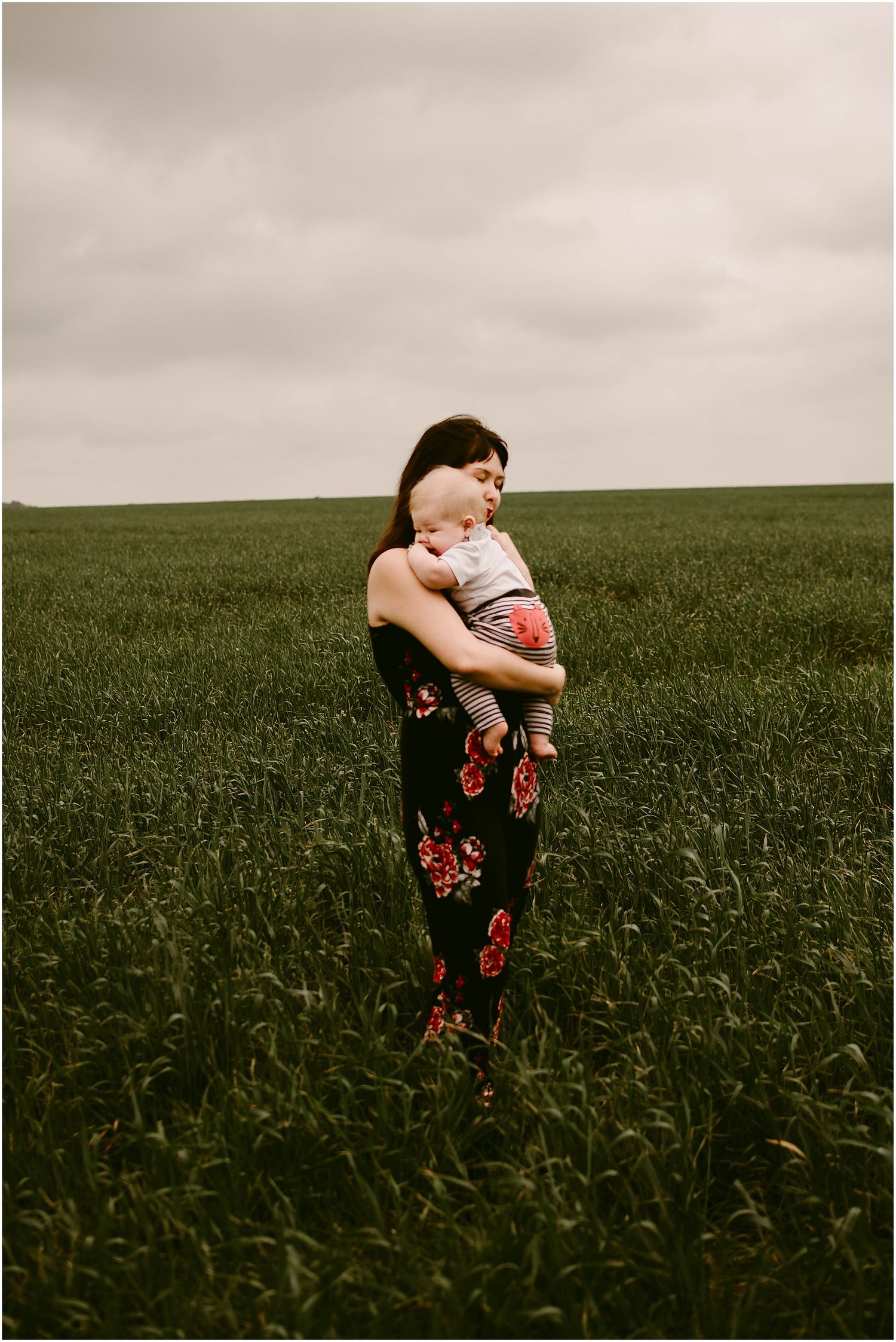 A woman in a dark floral dress holds a baby while standing in a lush green field.