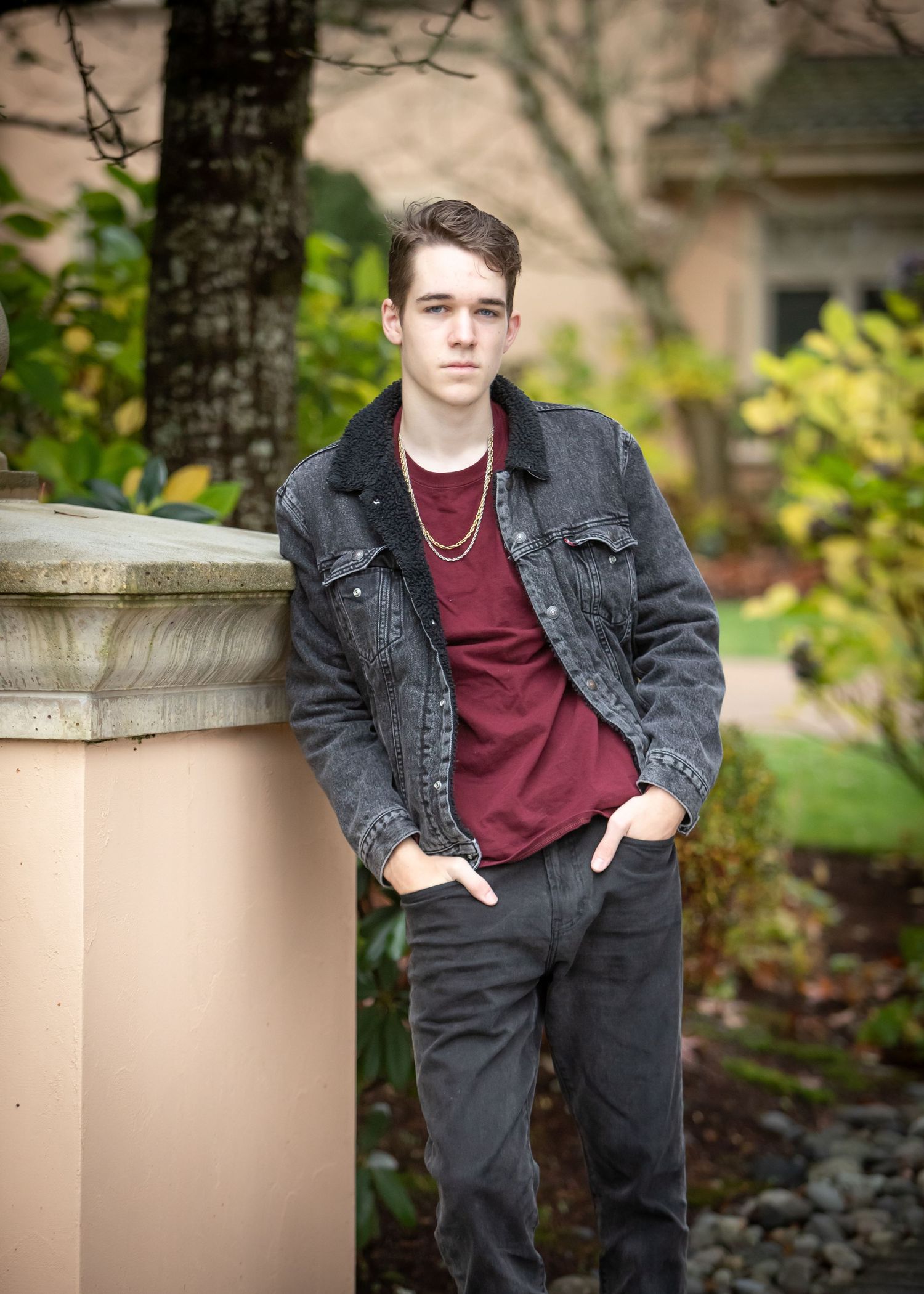 A casual portrait of someone in a burgundy shirt and denim jacket leaning against a stone wall.