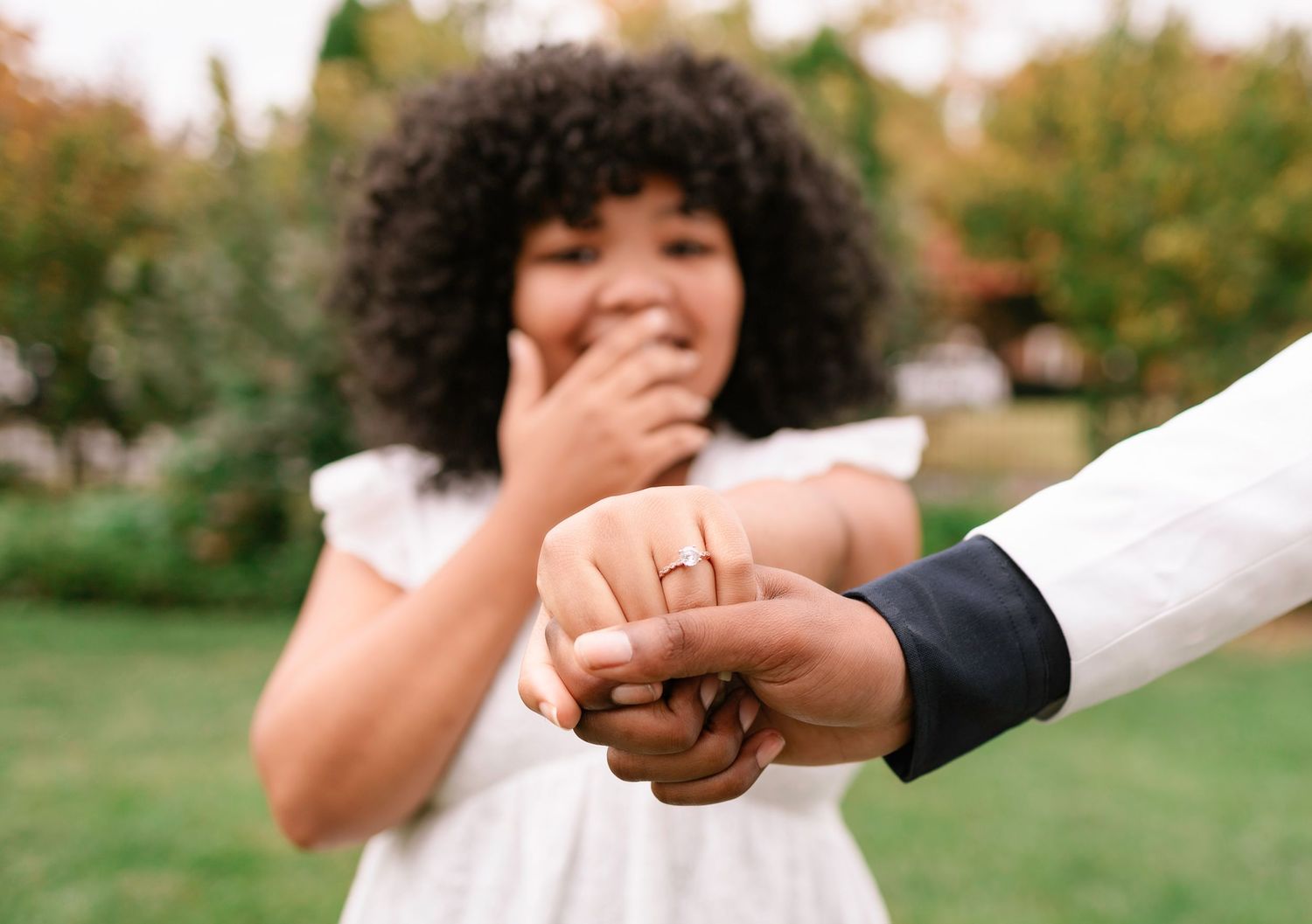 Woman with curly hair reacting with surprise while showing off her engagement ring outdoors in a garden setting.