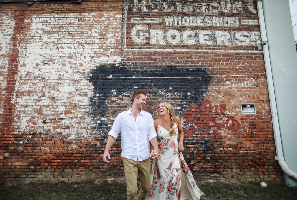 Couple stands against rustic brick wall with faded vintage grocers sign.