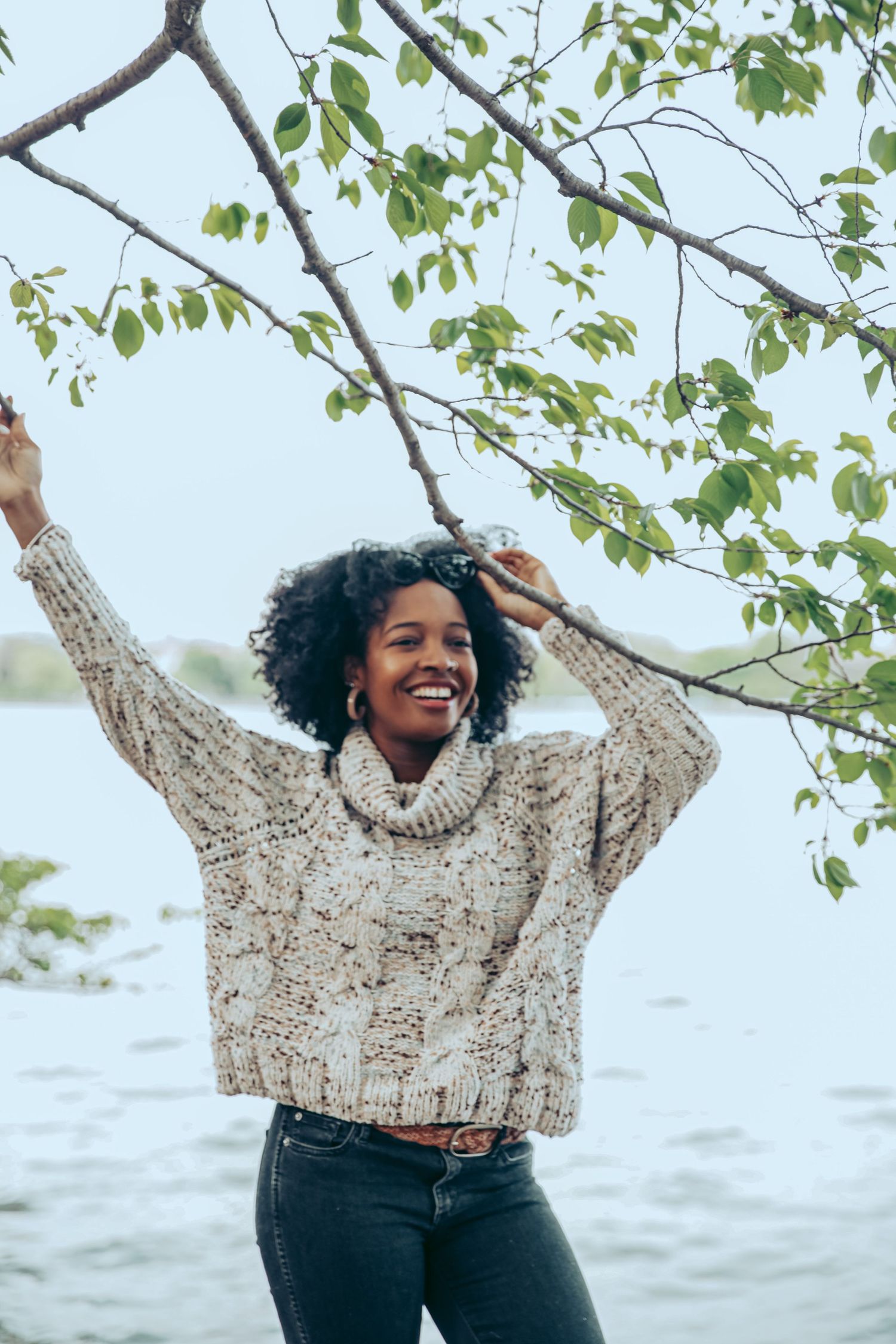 Smiling woman in cream cable-knit sweater reaching up to tree branches near lakeside.