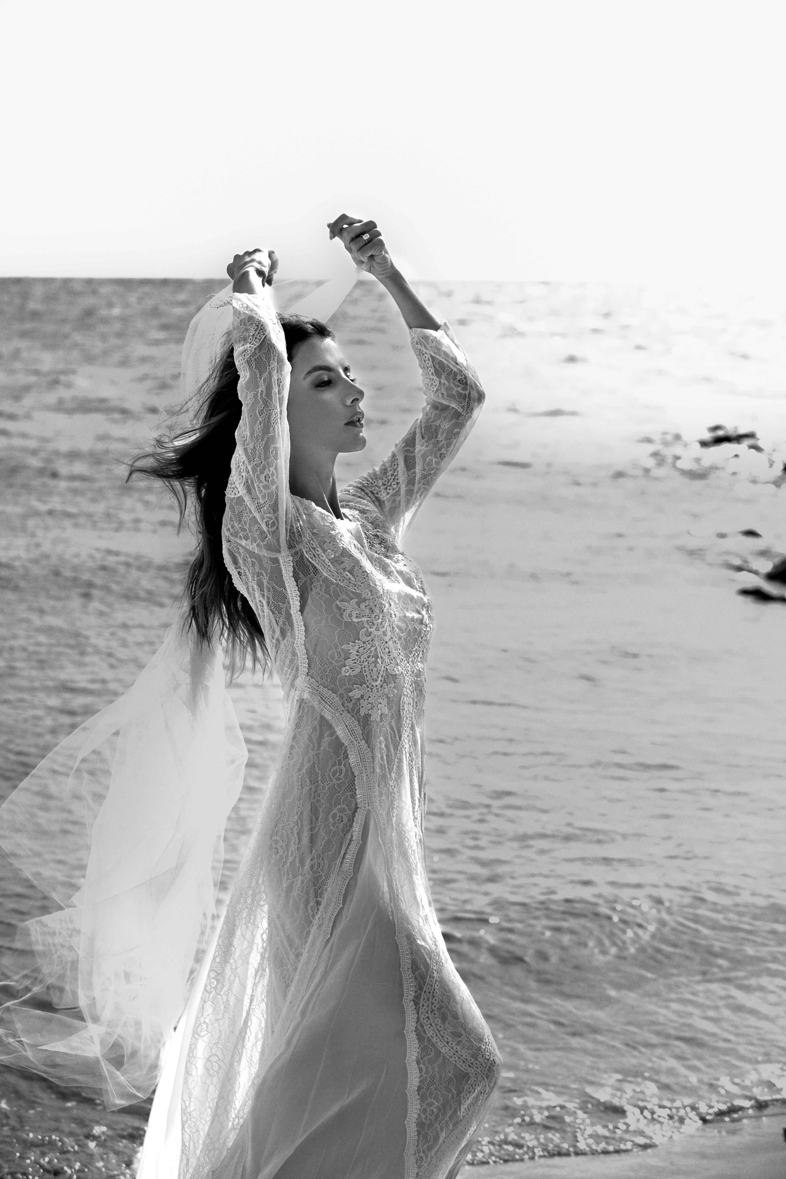 A person in a flowing white dress poses dramatically at the beach in black and white photography.
