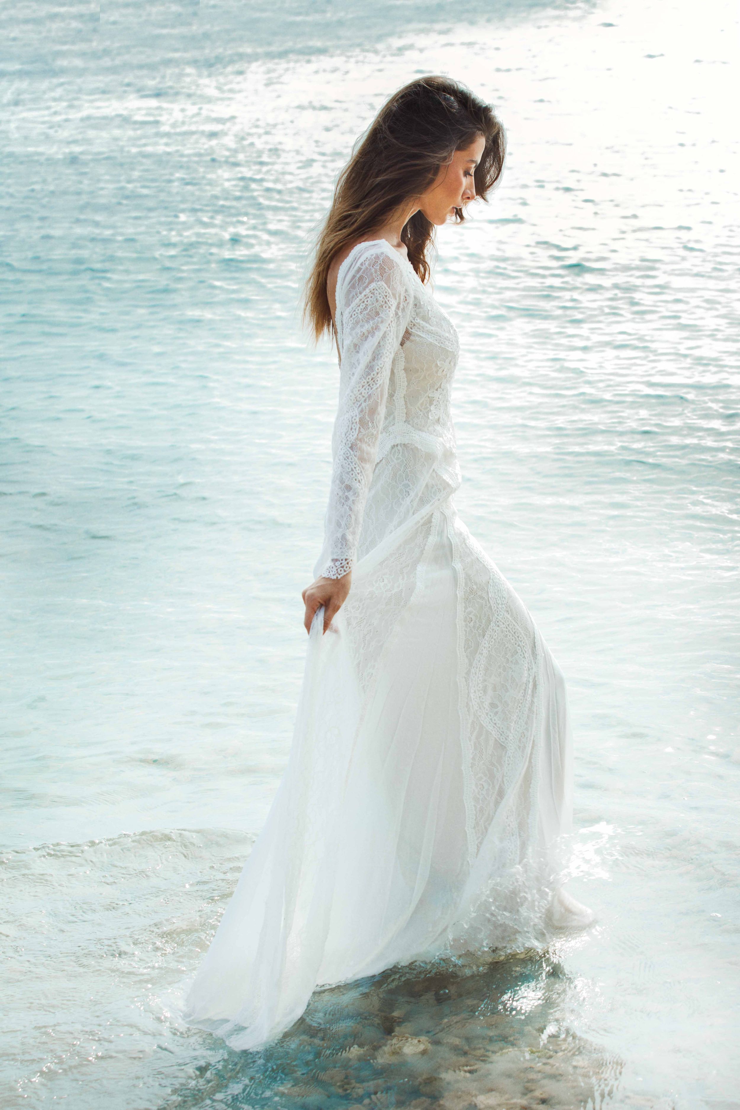 A figure in a flowing white lace dress stands at the edge of turquoise ocean waters on a bright sunny day.