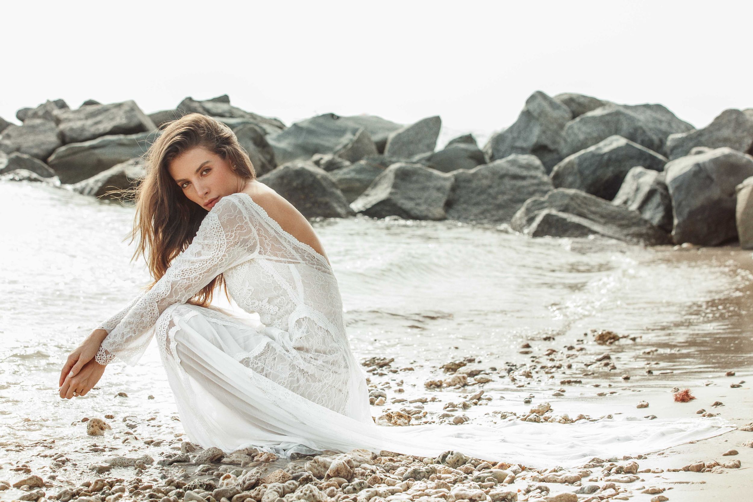 White flowy dress being worn for a romantic beach photoshoot at sunset near coastal rocks.