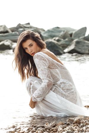 A woman in a flowing white lace dress poses elegantly on rocky beach terrain at sunset.