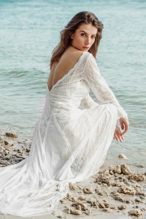 Elegant white lace wedding dress with long sleeves photographed against turquoise ocean waters on rocky beach.