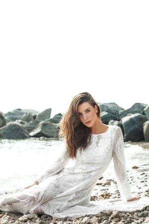 Woman in flowing white lace dress poses gracefully on rocky beach against ocean backdrop.