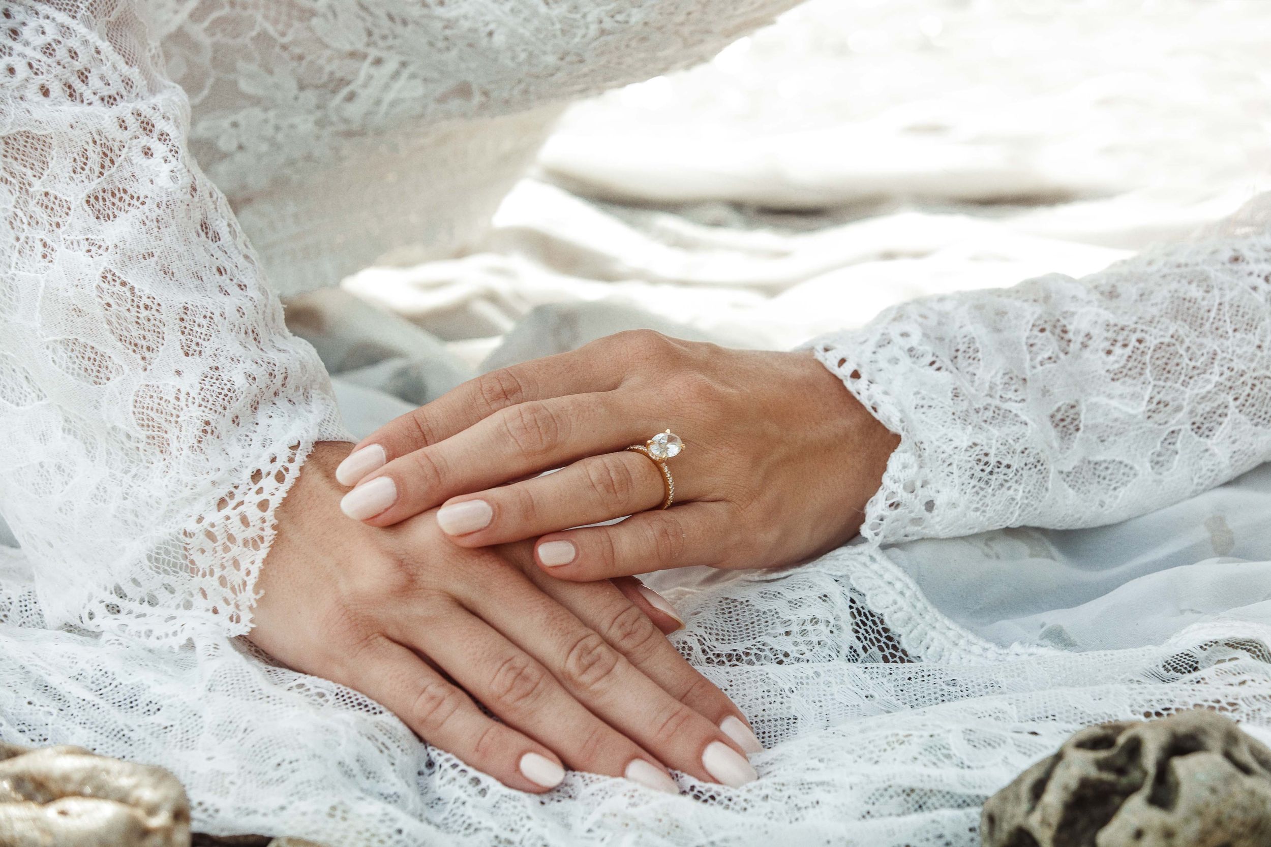 Close-up of hands wearing a white lace dress sleeve and elegant diamond engagement ring on light fabric background.