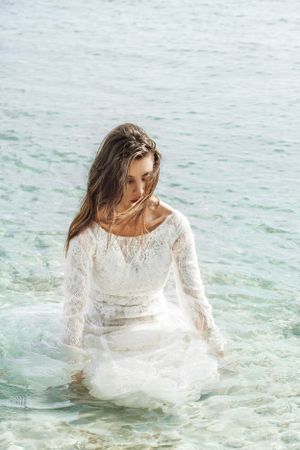 A person in a flowing white lace dress poses ethereally in crystal clear turquoise ocean waters.