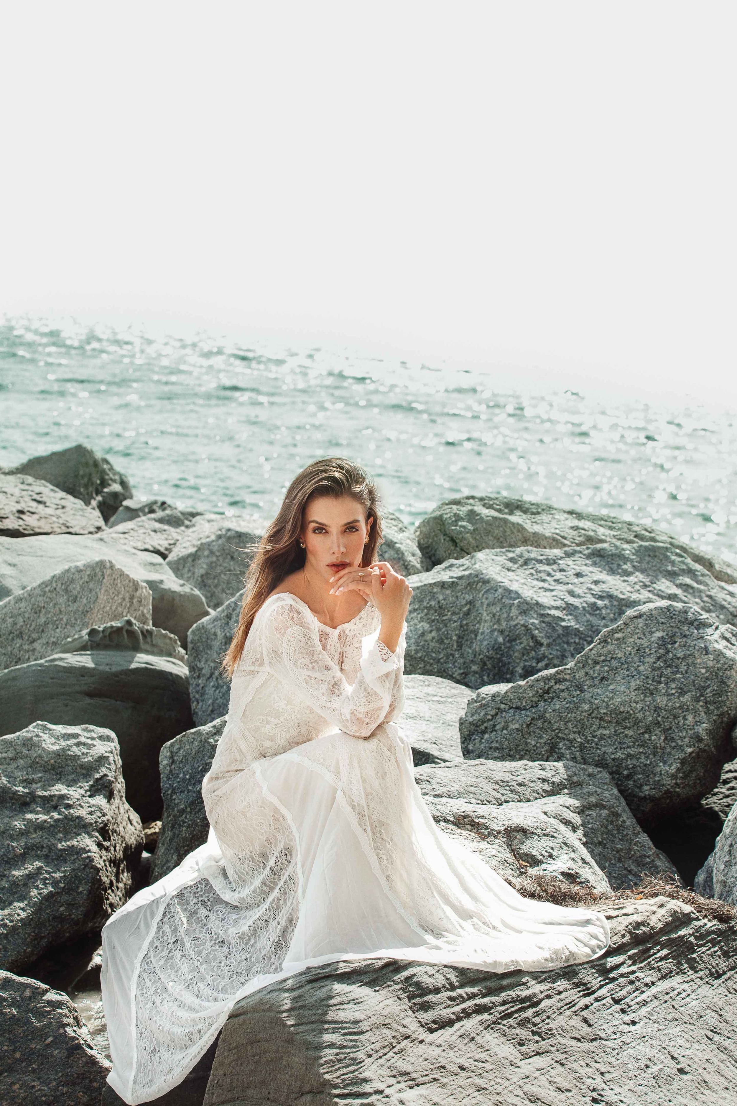 Woman in flowing white lace dress poses elegantly on coastal rocks with turquoise ocean waves in background.