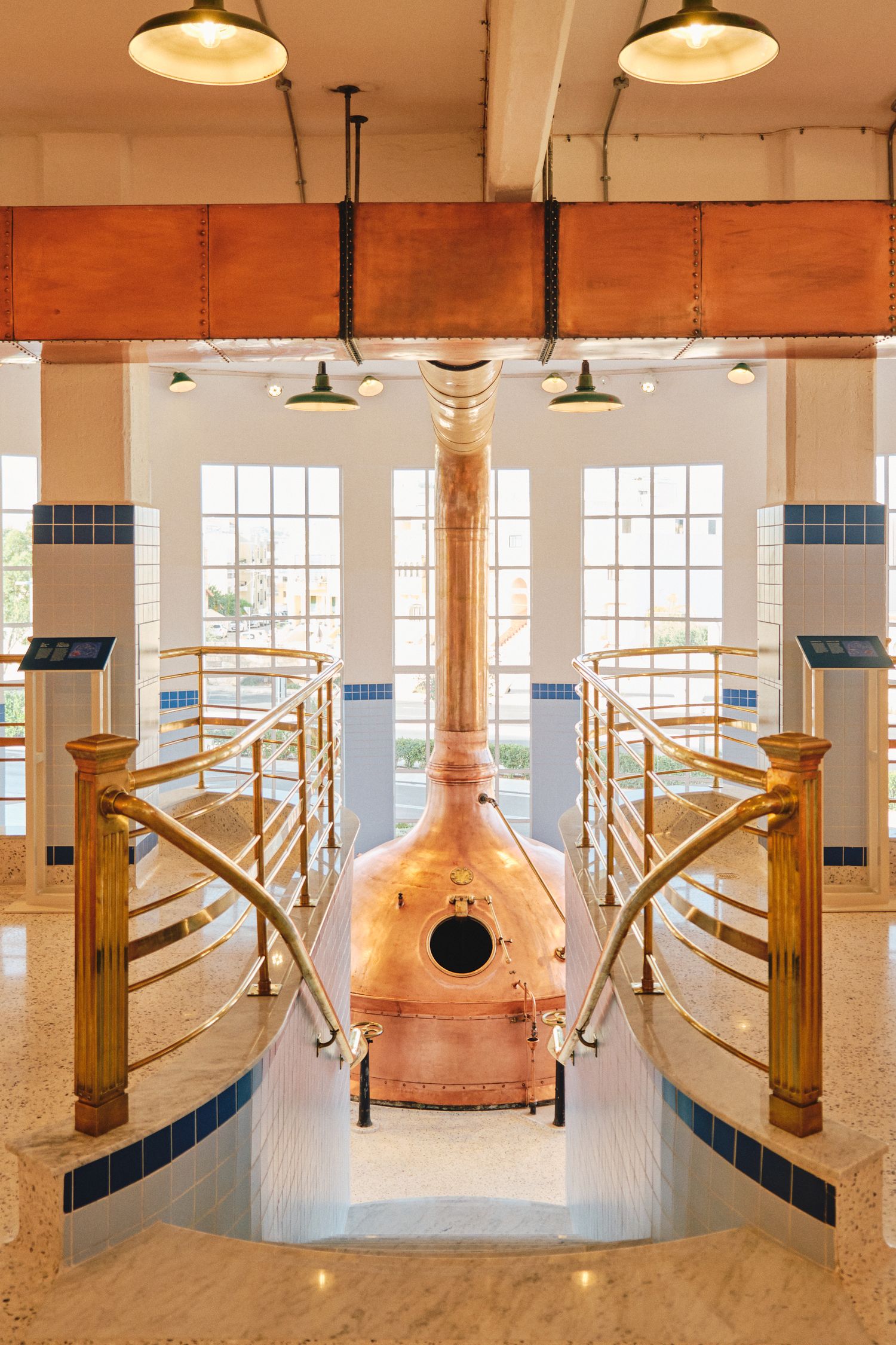 Large copper brewing kettle in an elegant white-tiled brewery room with wooden railings.