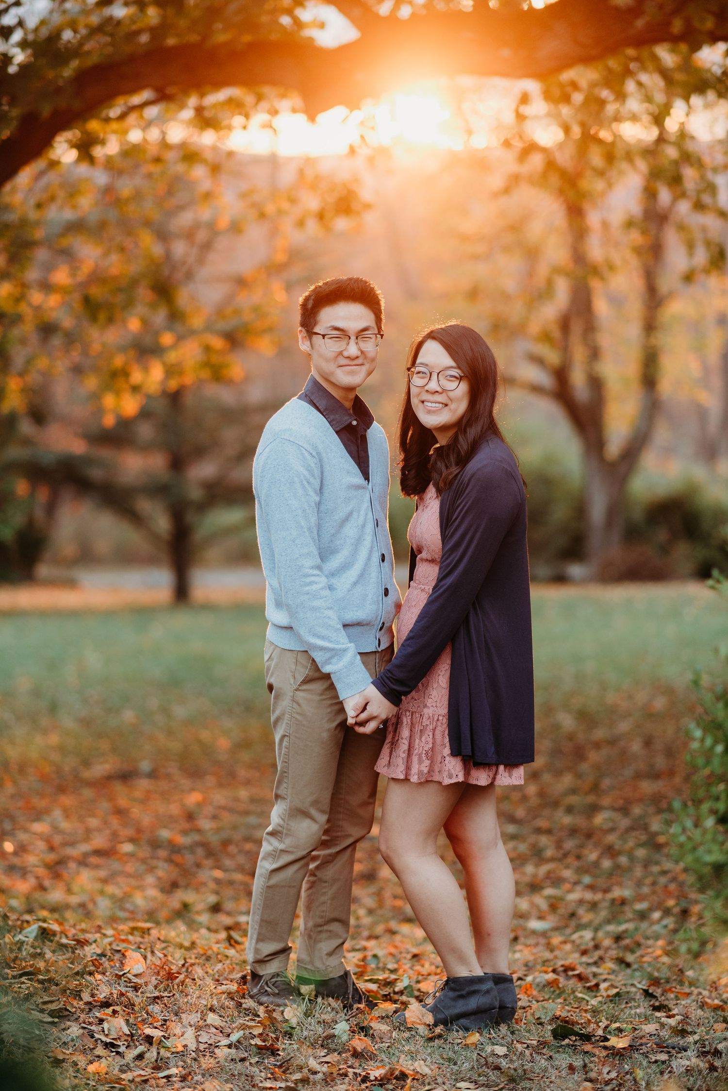 A couple stands together on fallen autumn leaves during golden hour sunset in a park setting at Ringwood Manor.