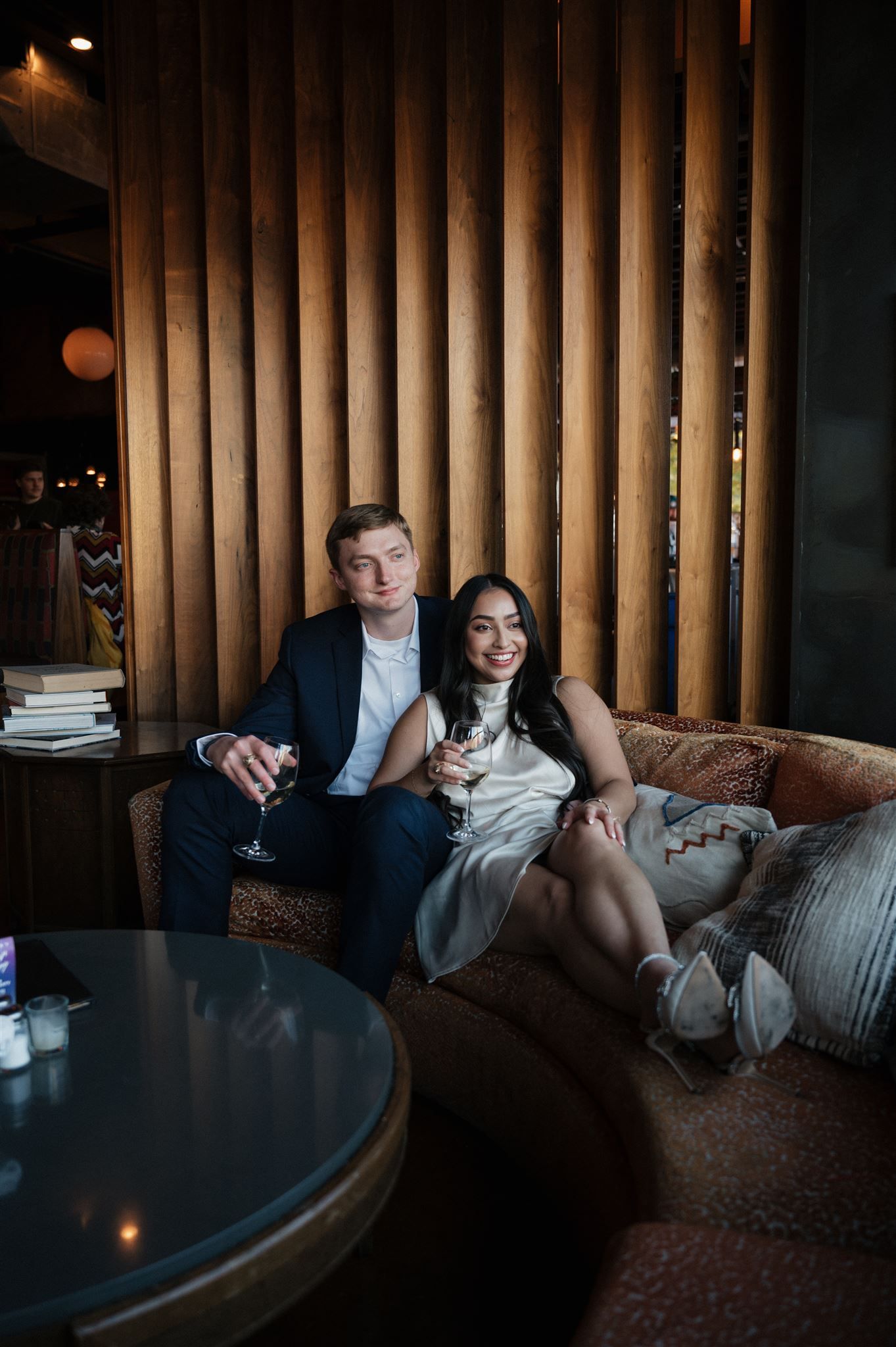 Couple lounging on leather couch sharing wine in atmospheric restaurant with wooden wall panels.