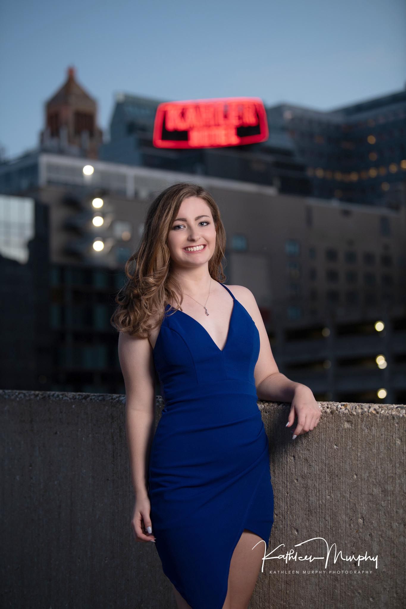 Evening portrait in a navy blue dress with illuminated city buildings in background.