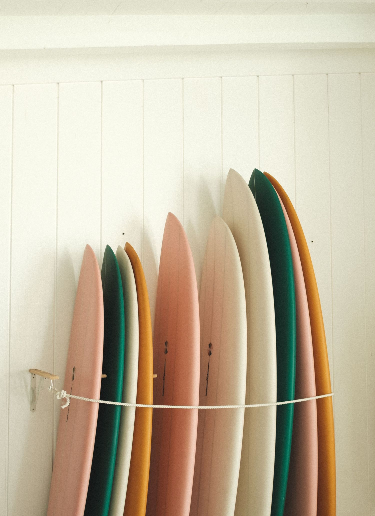 Collection of surfboards in pastel and earth tones lined up against a white wall.