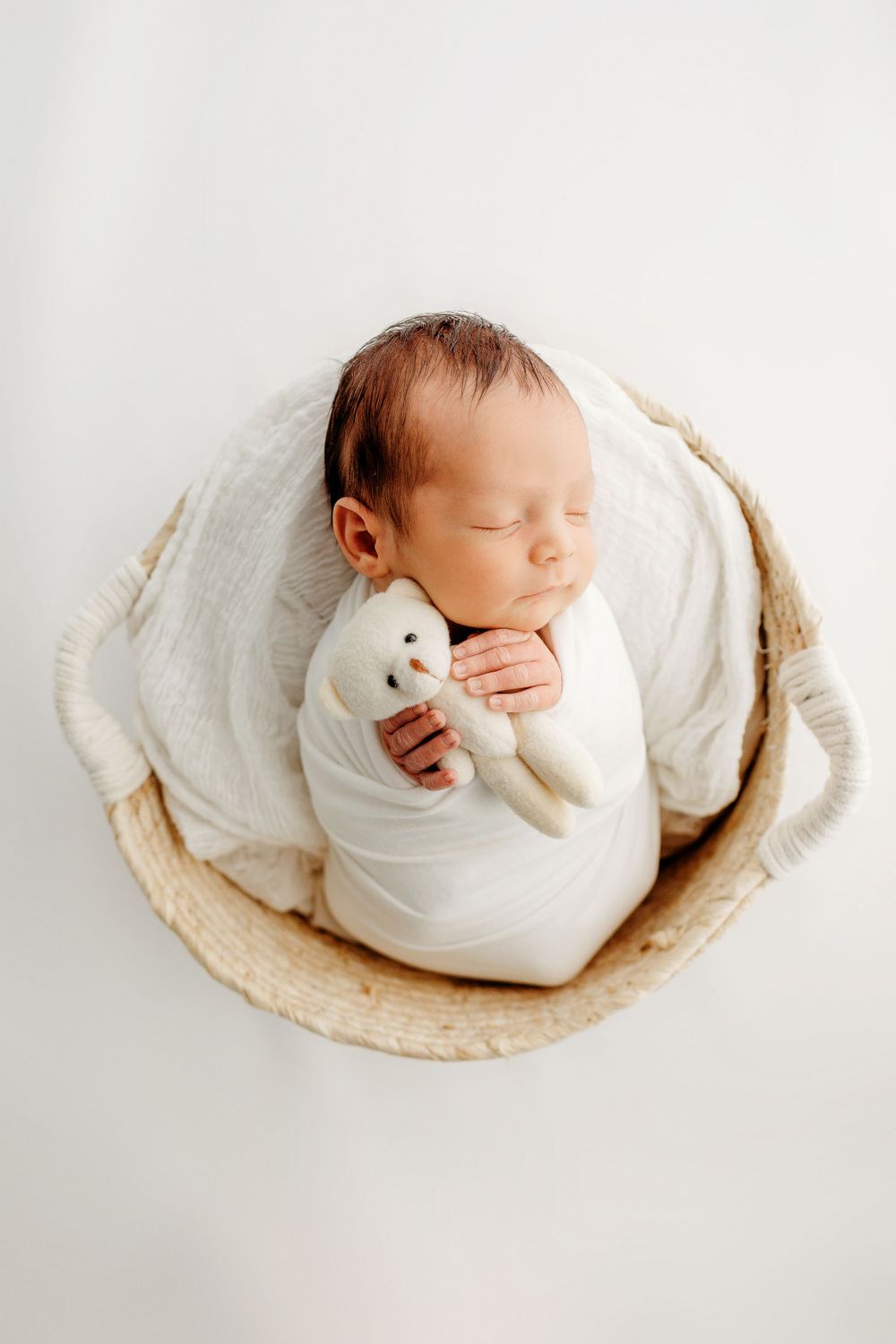 Peaceful newborn sleeping in round basket with white wrap and plush toy.