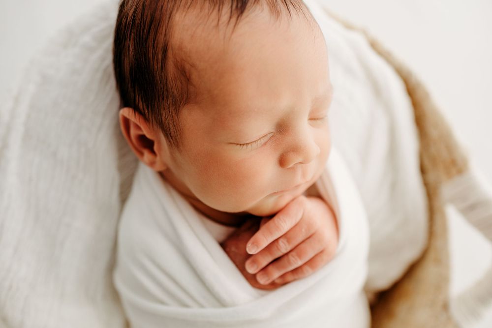 Peaceful sleeping newborn swaddled in soft white blanket against neutral background.