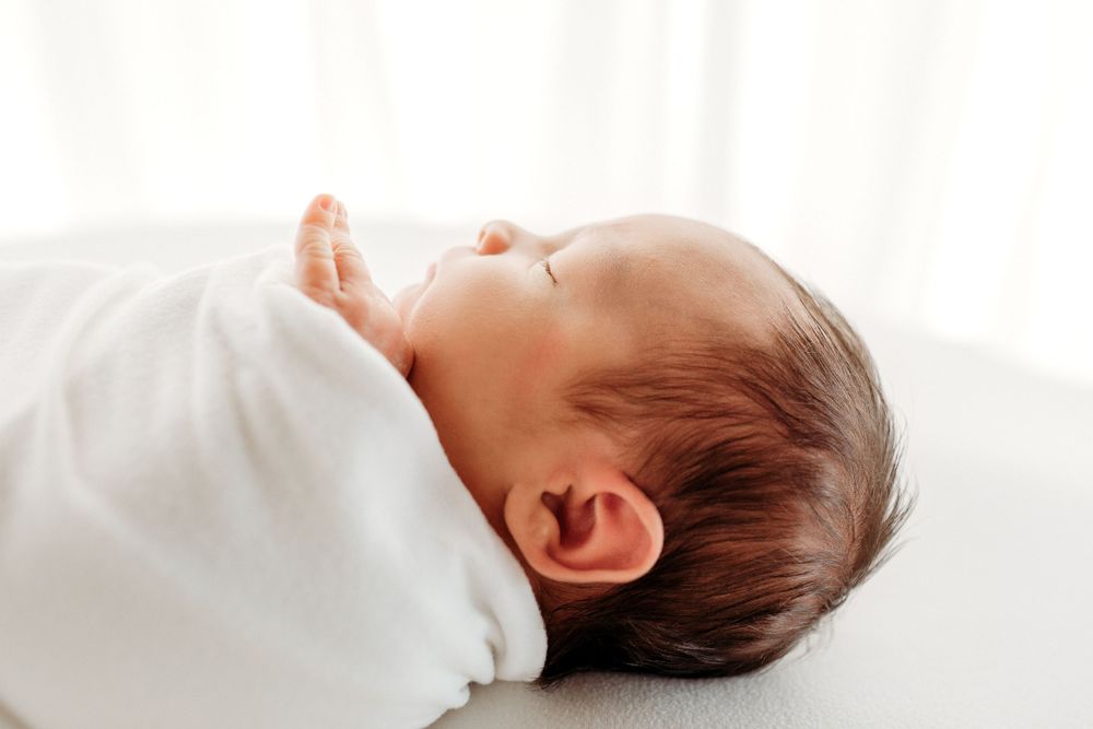 Side view of sleeping newborn in soft white wrap against bright background.