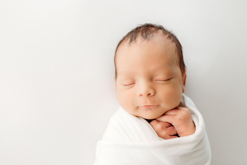 Newborn baby wrapped in white swaddle sleeping peacefully on light background.