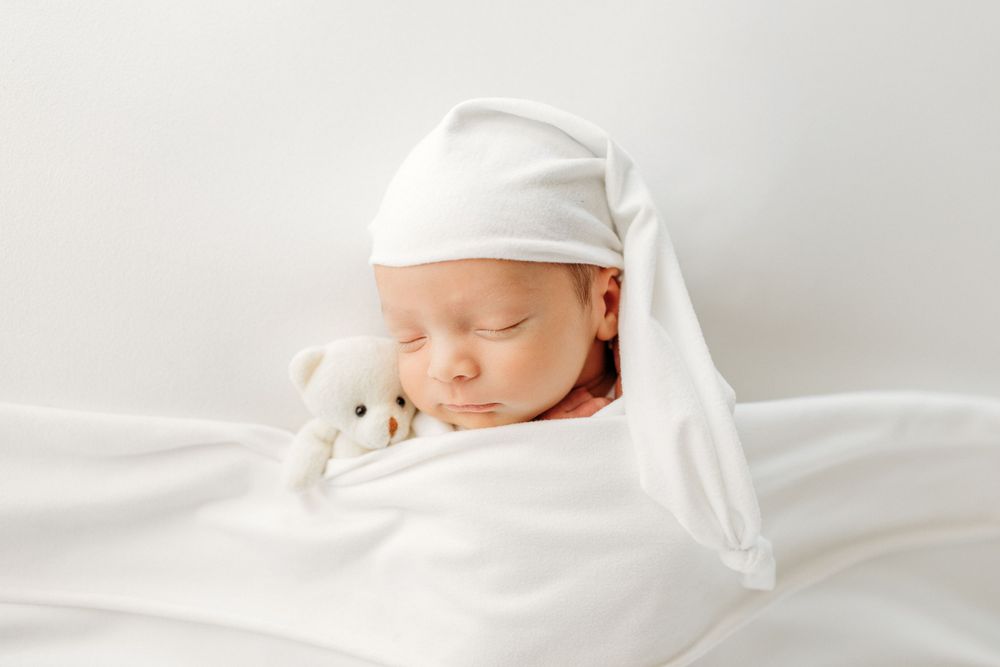 Sleeping baby wearing white cap cuddled with small stuffed toy.