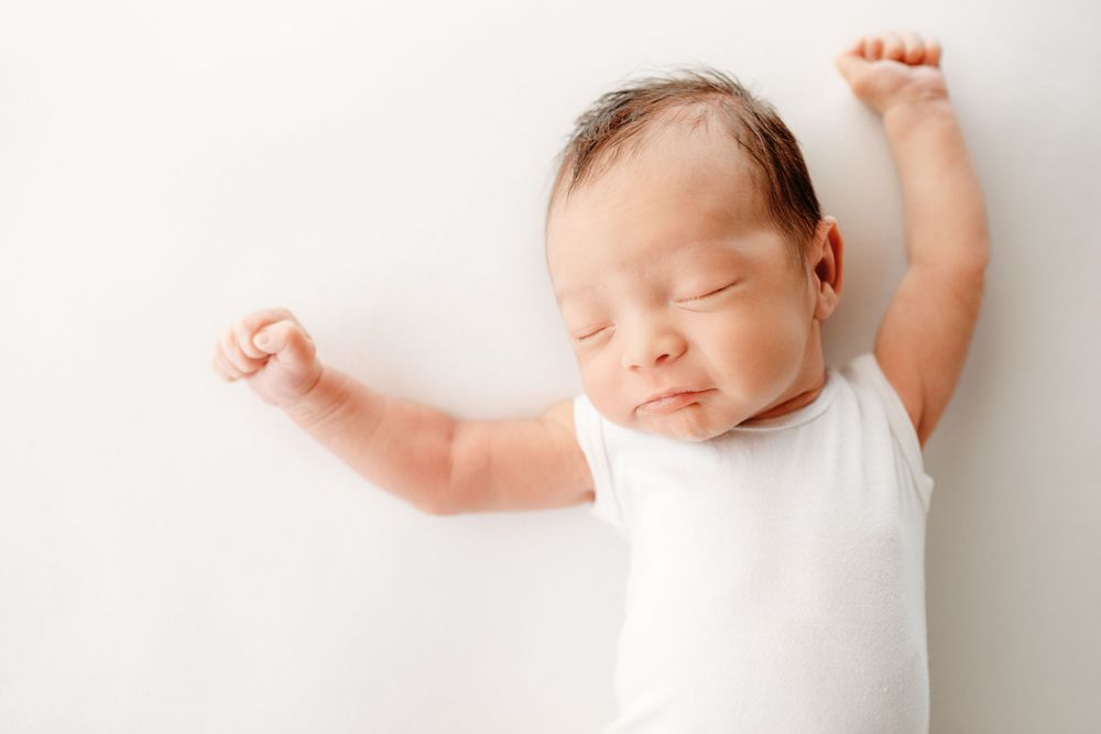 Sleeping newborn in white onesie stretching arms on white background.