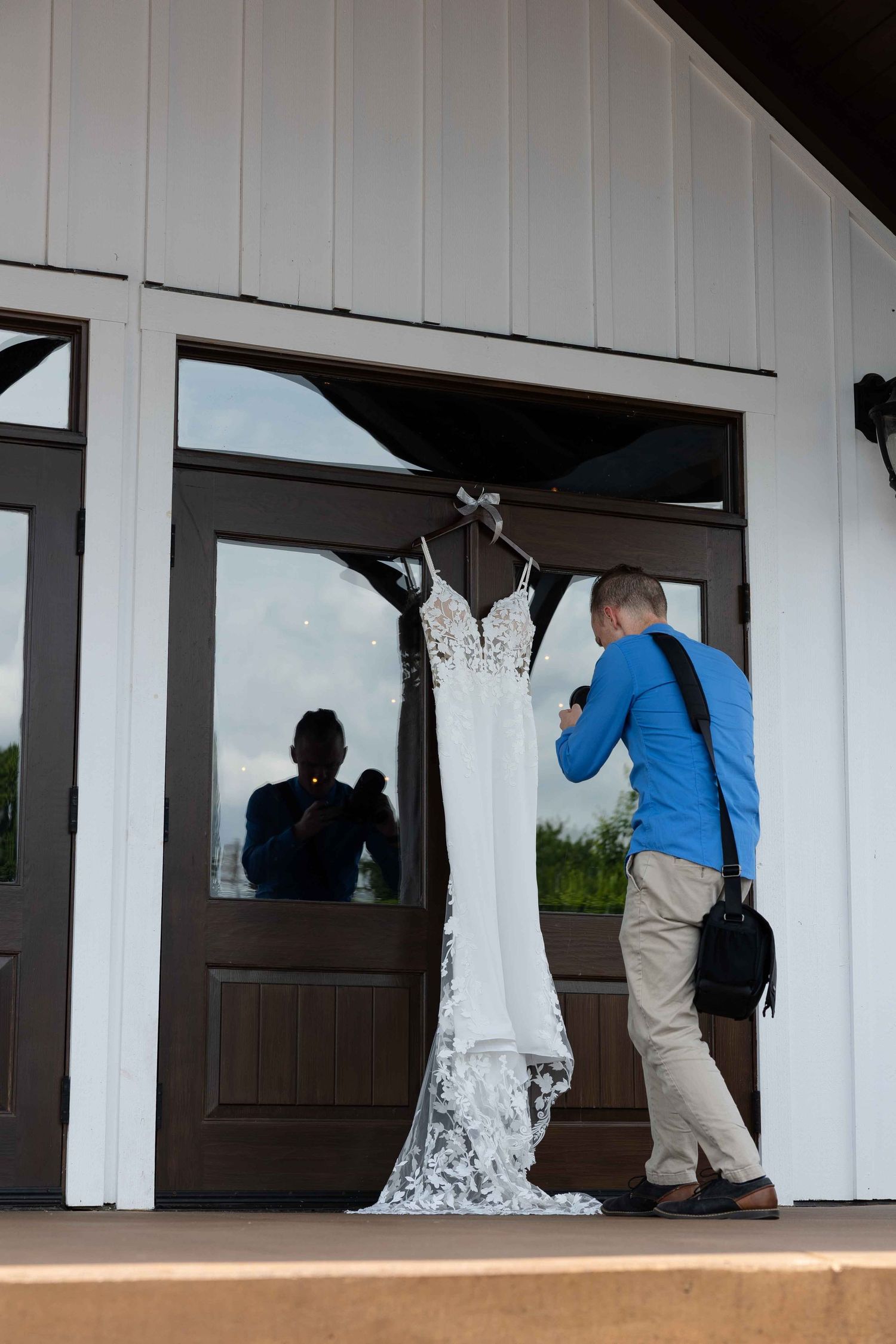 Wedding dress hanging outside white barn building while photographer captures detail shots.
